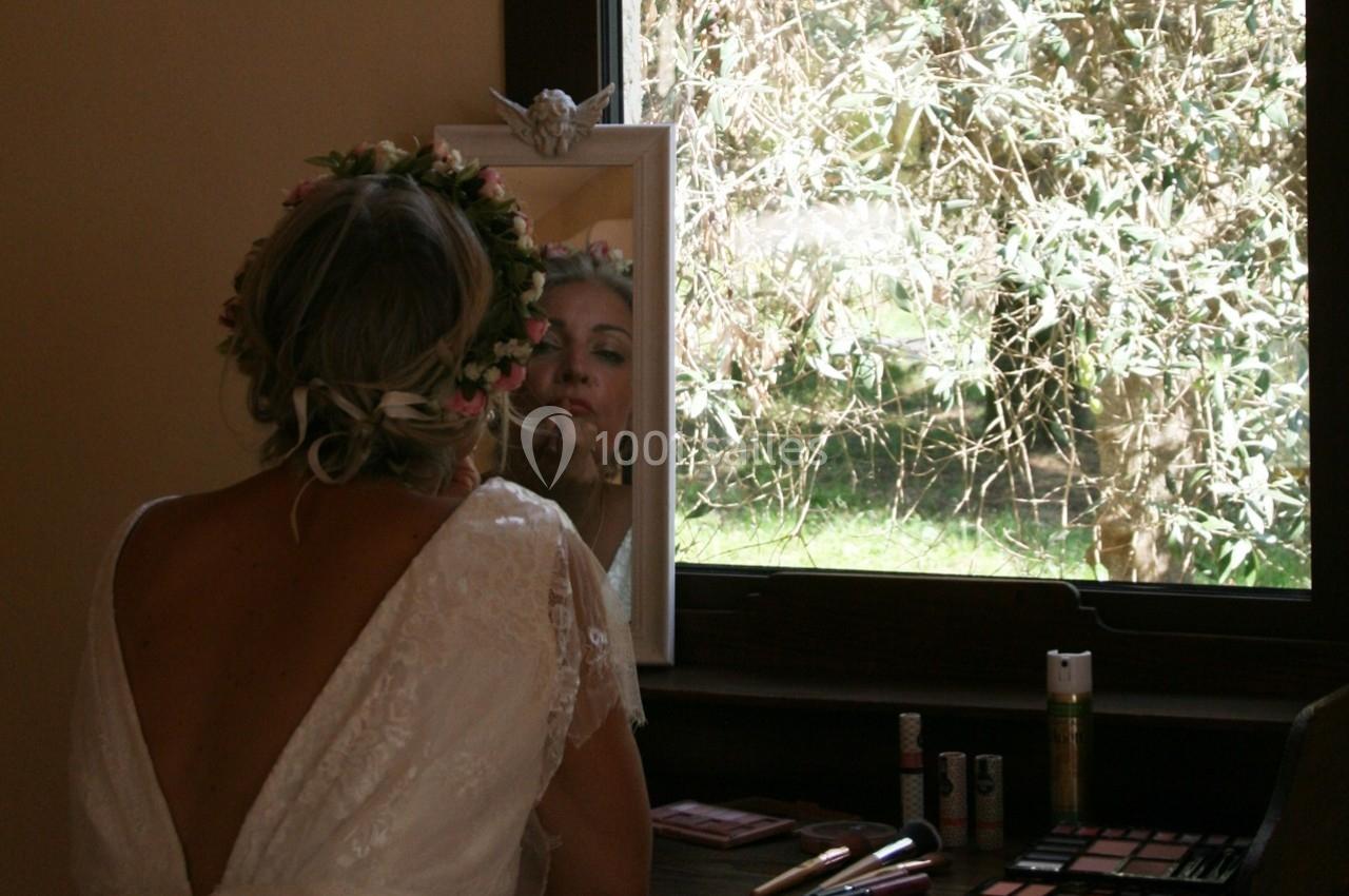 Une femme en robe blanche et coiffée de fleurs se regarde dans un miroir près d'une fenêtre, entourée de maquillage.