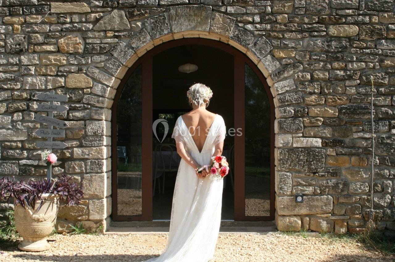 Une femme en robe de mariée blanche, vue de dos, tenant un bouquet devant une porte en pierre voûtée.