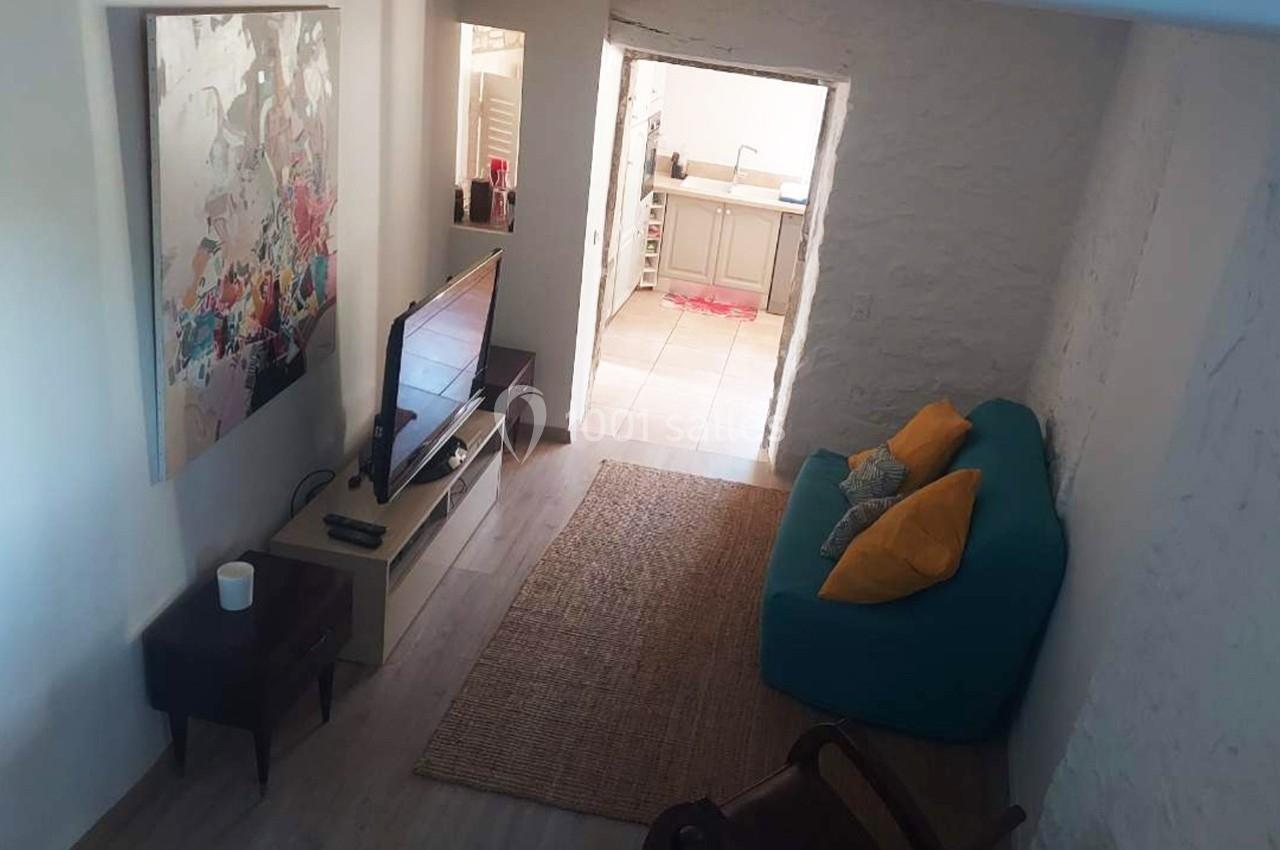 Salon lumineux avec canapé turquoise, coussins colorés, télévision, tapis en fibres naturelles et vue sur une cuisine…