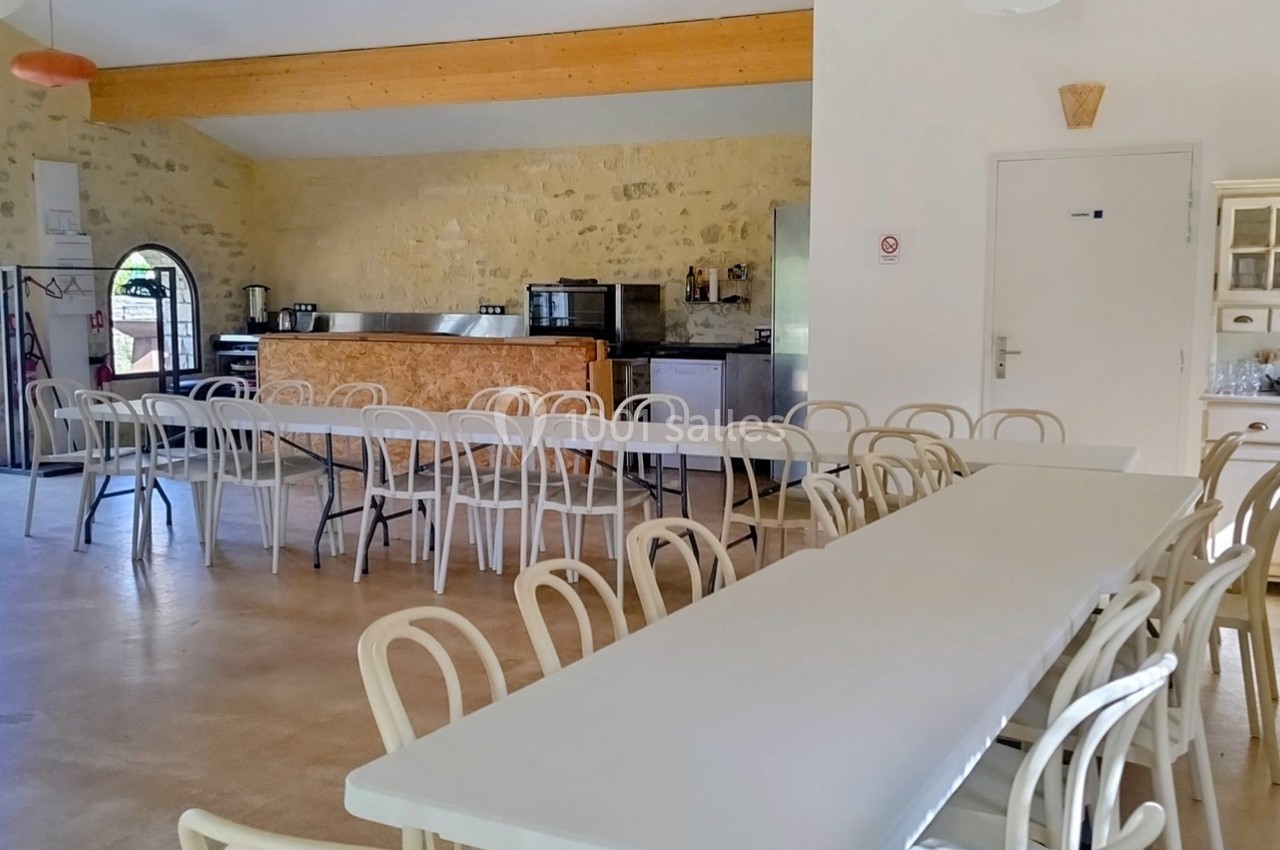 Salle lumineuse avec tables et chaises blanches, murs en pierre, cuisine ouverte et grandes portes vitrées donnant sur l…
