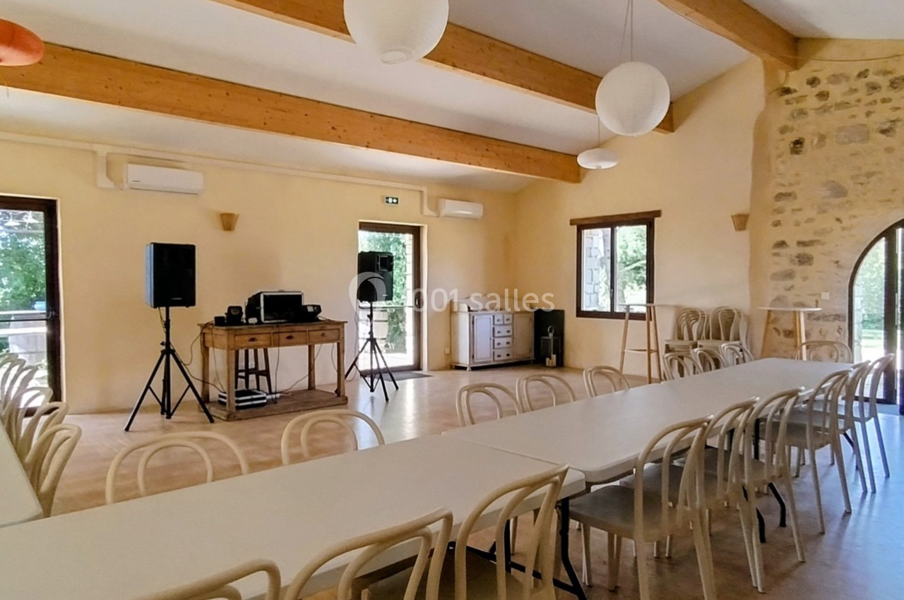 Salle lumineuse avec tables alignées, chaises en bois, murs en pierre et équipements audio près des fenêtres.