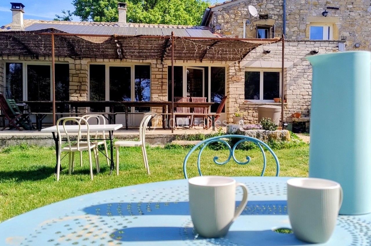 Table en métal avec deux tasses et une carafe bleue, devant une maison en pierre avec terrasse ombragée.