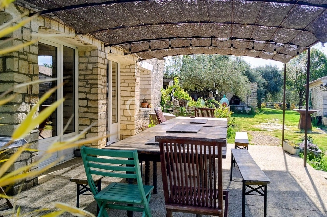 Terrasse ombragée avec table en bois et chaises, entourée de pierres et de verdure dans un jardin ensoleillé.