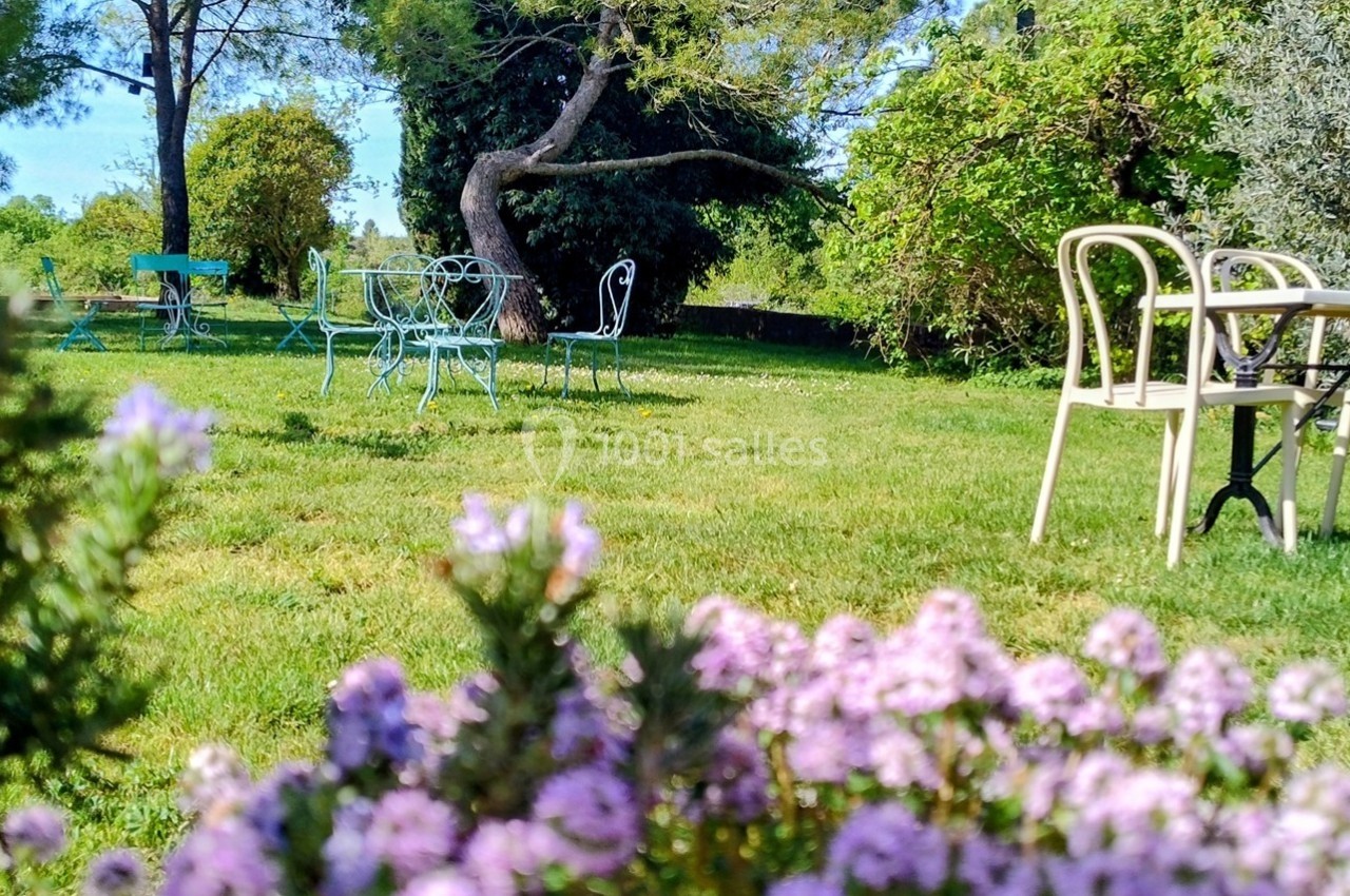 Chaises et tables en métal disposées dans un jardin verdoyant avec des fleurs violettes au premier plan.