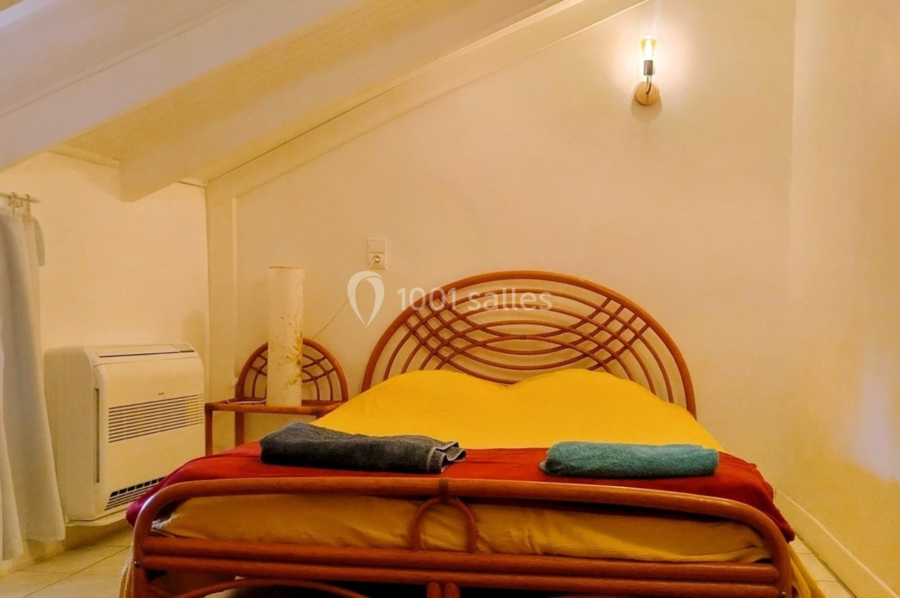 Chambre mansardée avec lit double en rotin, linge plié, murs clairs et petite fenêtre avec rideaux blancs.