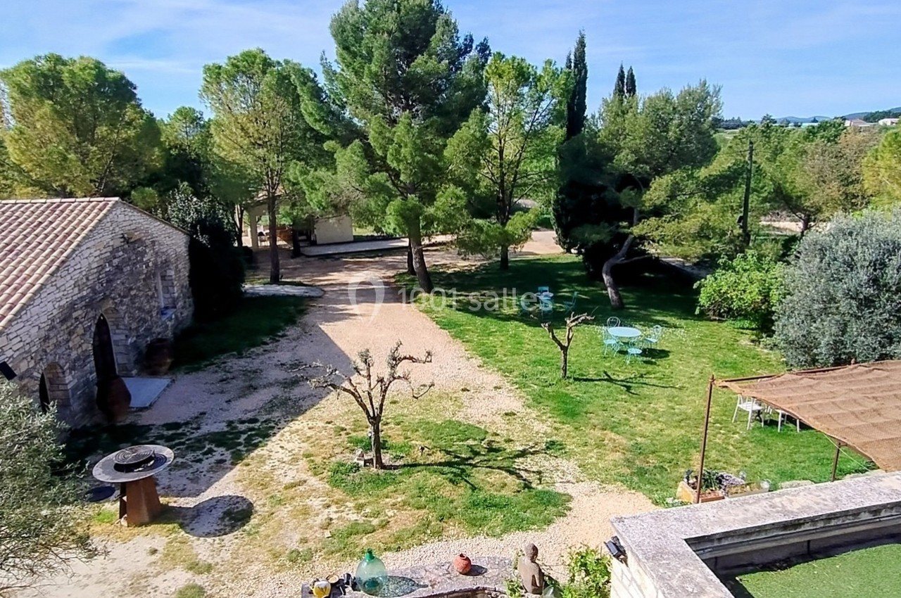 Vue d'un jardin arboré avec une terrasse, des tables, un puits et des bâtiments en pierre sous un ciel dégagé.