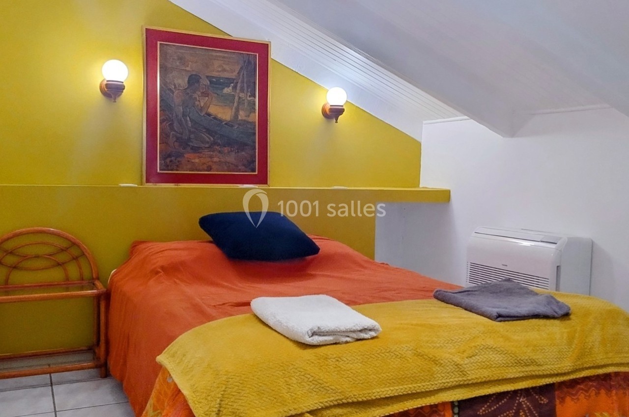 Chambre mansardée avec lit double, mur jaune, tableau encadré, deux appliques murales et linge de lit coloré.