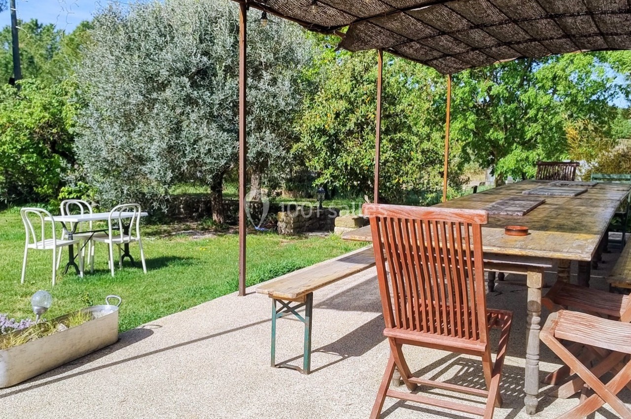 Terrasse ombragée avec une grande table en bois, entourée de chaises, donnant sur un jardin verdoyant.