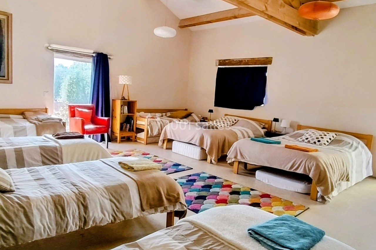 Chambre lumineuse avec plusieurs lits simples, tapis coloré au sol et poutres apparentes au plafond.