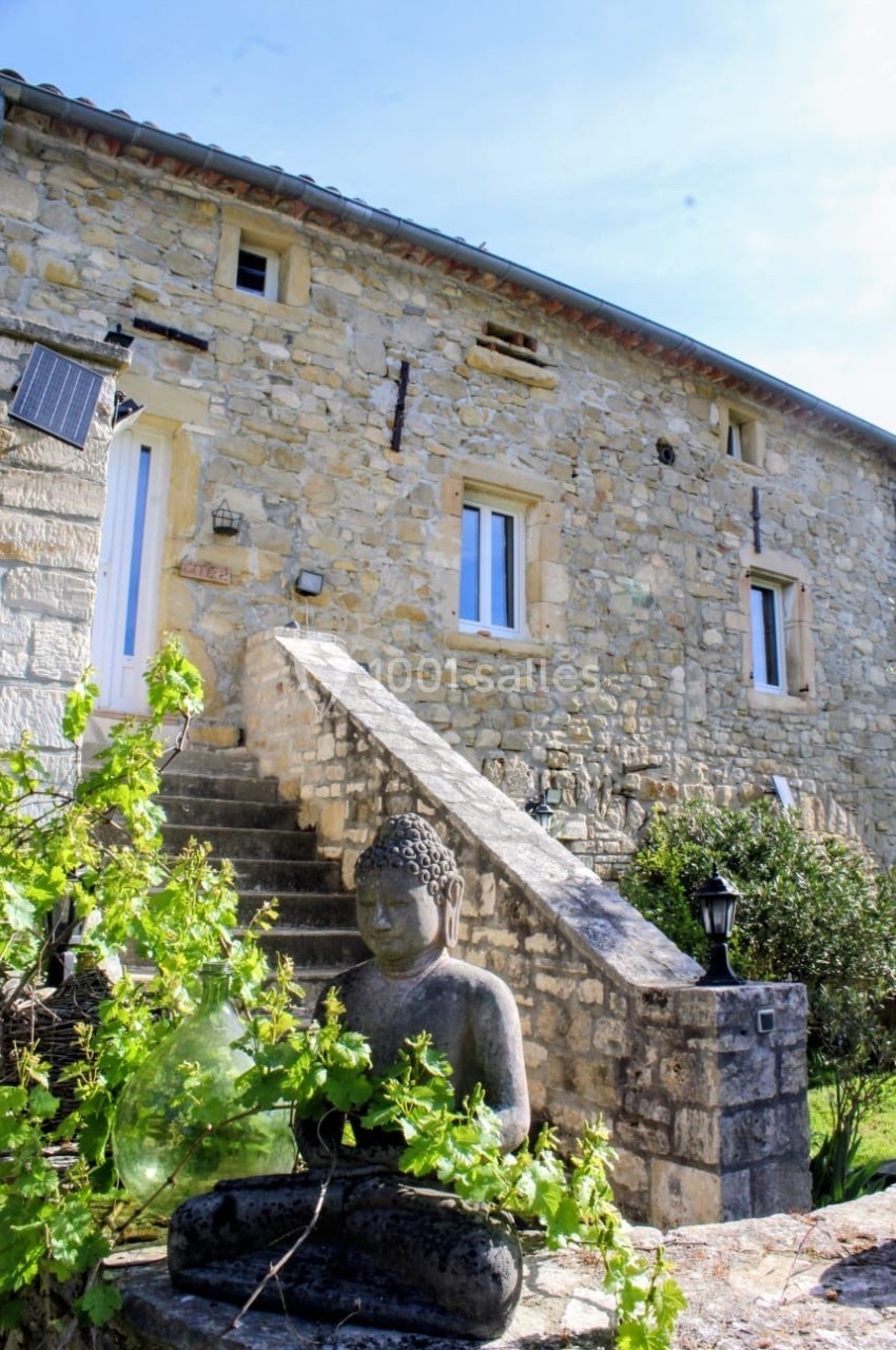 Statue de Bouddha en pierre devant une maison en pierre avec escalier et végétation environnante.