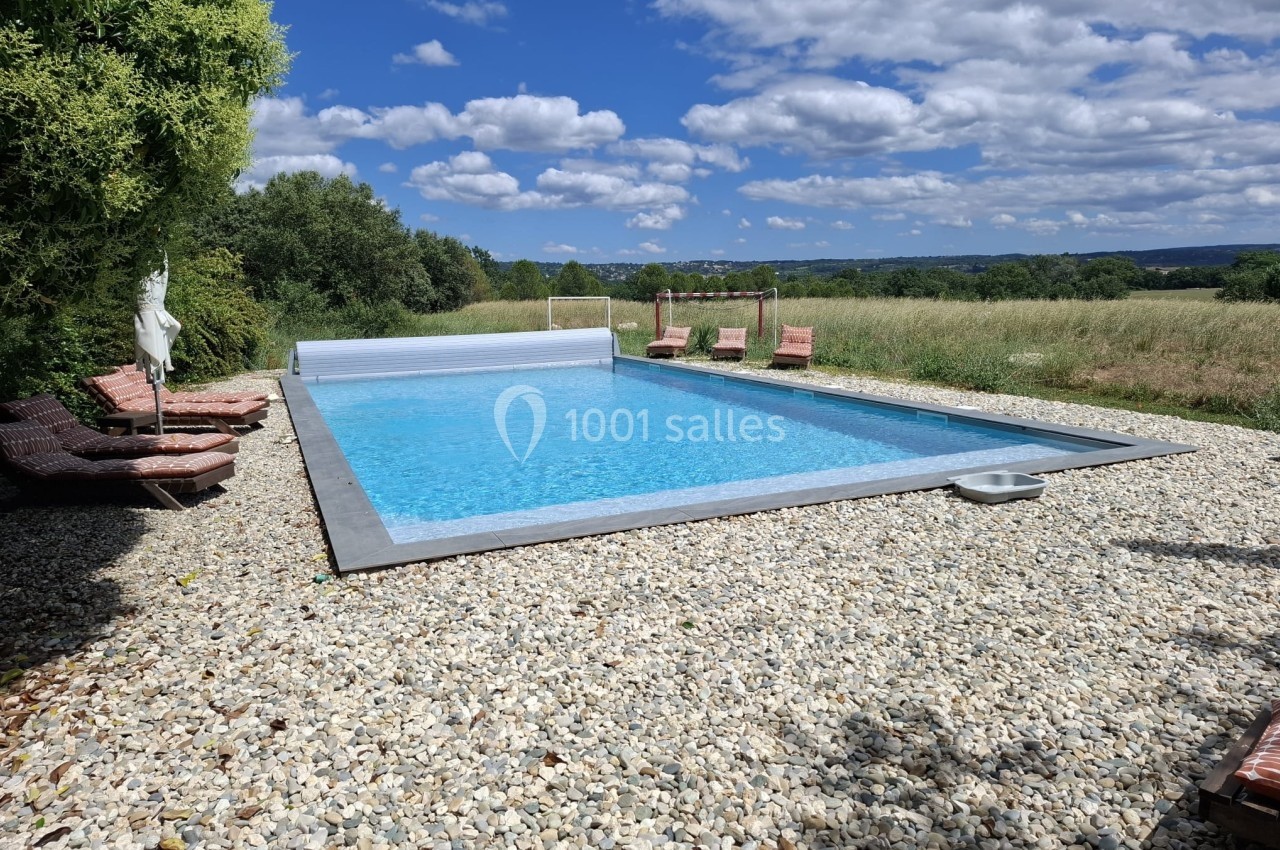 Piscine extérieure entourée de galets, avec chaises longues et vue sur un paysage champêtre sous un ciel bleu.