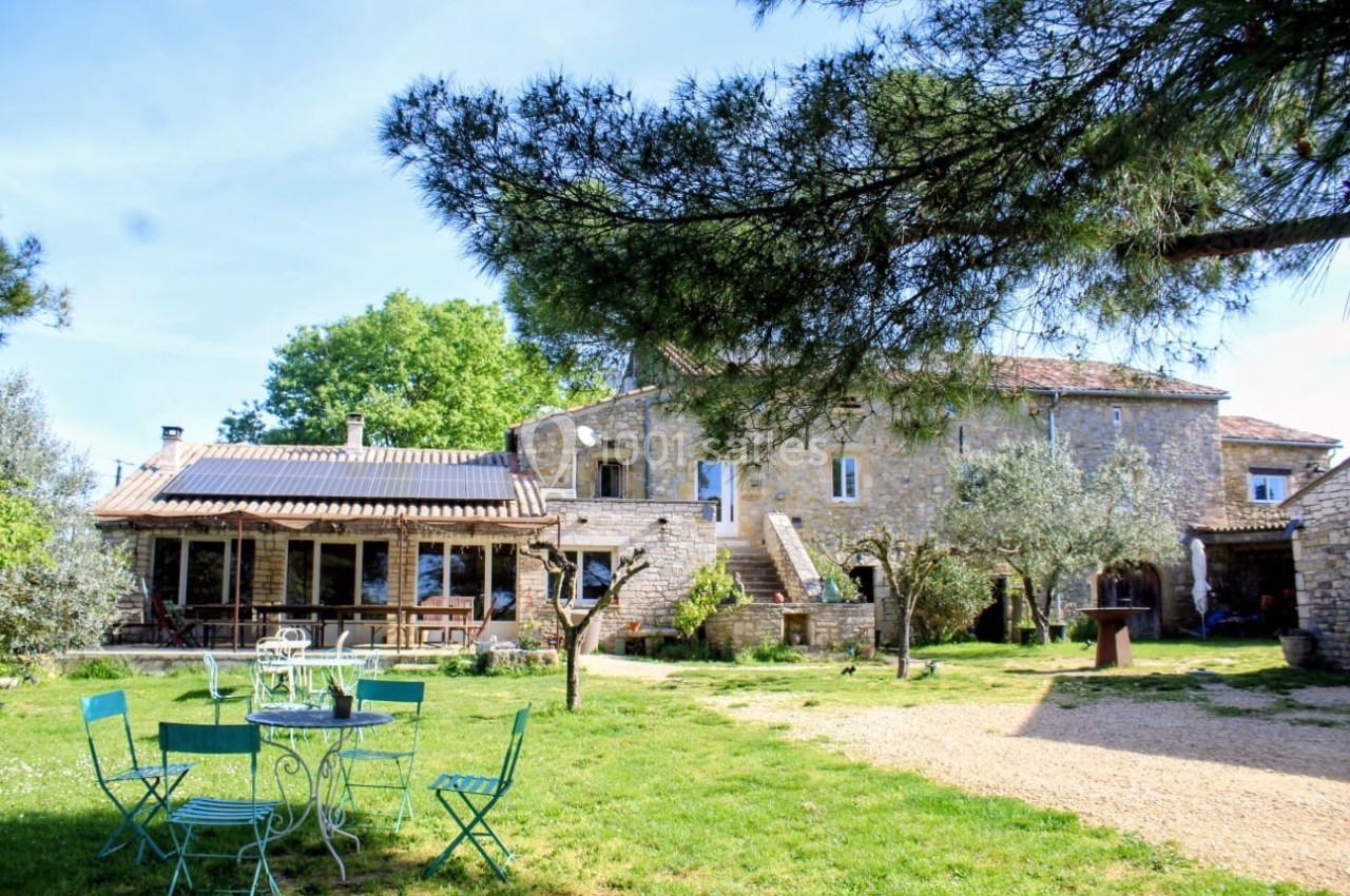 Cour d'une maison en pierre avec mobilier de jardin vert, arbres et pelouse sous un ciel dégagé.