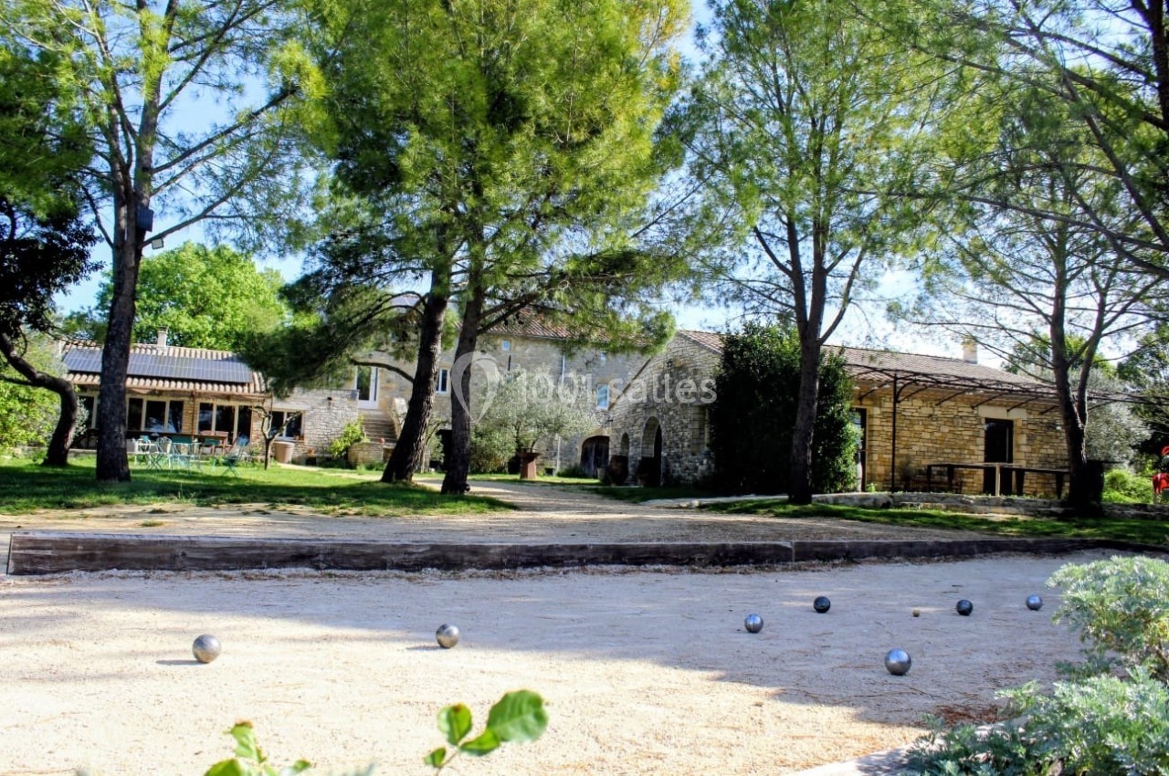 Terrain de pétanque ombragé devant des bâtiments en pierre entourés d'arbres et de végétation.