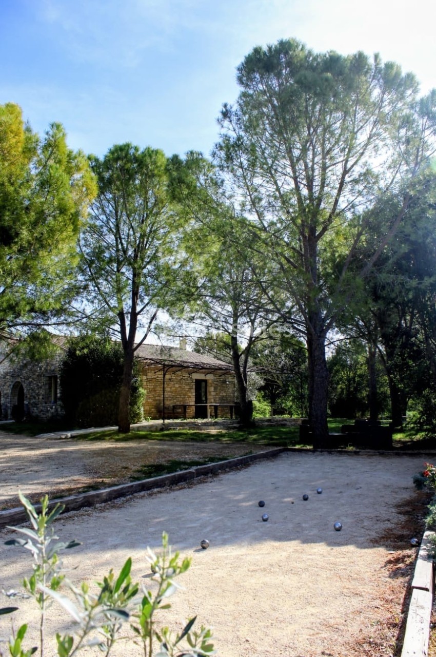 Terrain de pétanque ombragé entouré d'arbres et d'un bâtiment en pierre sous un ciel dégagé.