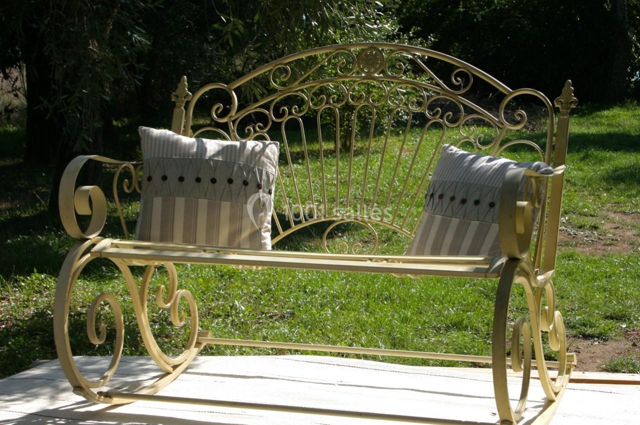 Banc en fer forgé jaune avec deux coussins rayés, placé sur une terrasse en bois dans un jardin verdoyant.