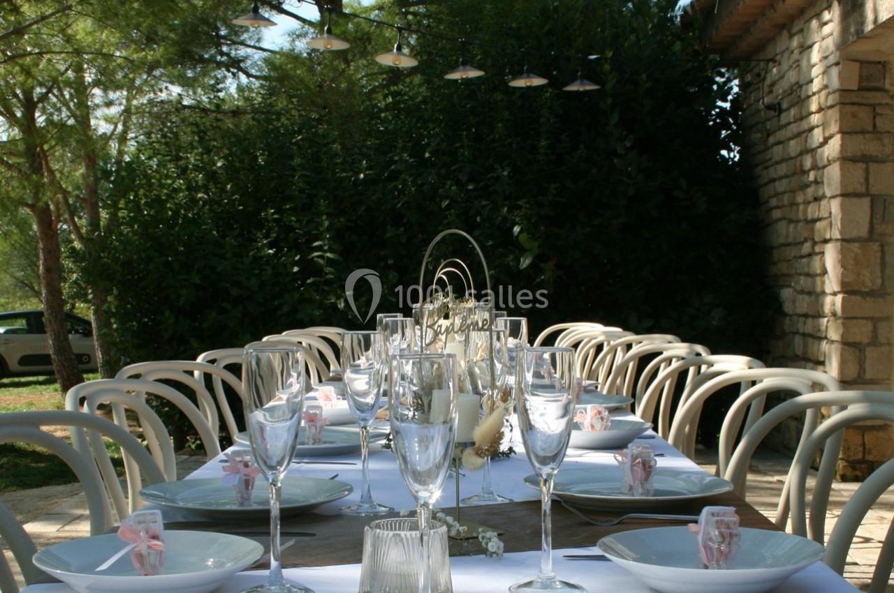 Table extérieure dressée avec vaisselle blanche, verres à pied et décorations, entourée de verdure et éclairée par des…