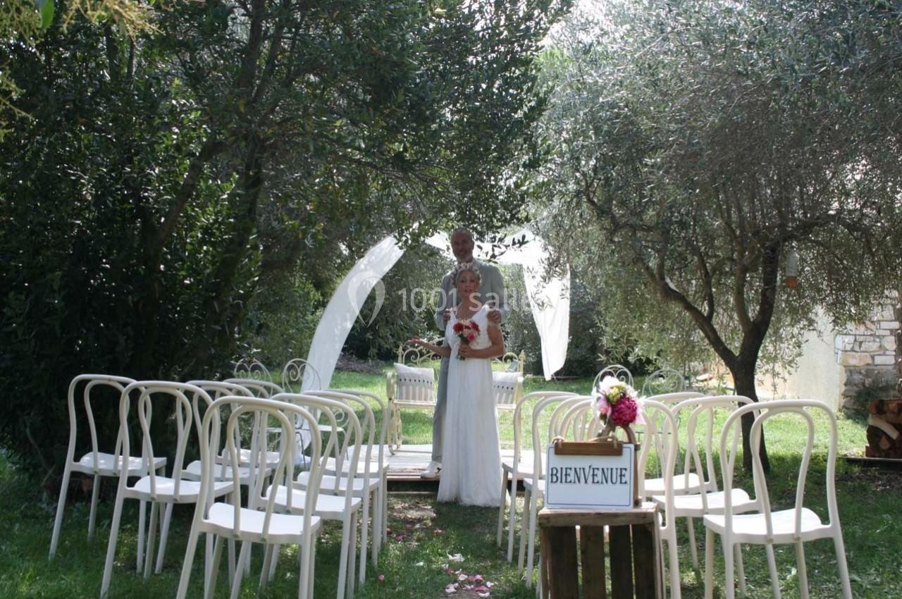 Un couple de mariés pose dans une allée bordée de chaises blanches, sous des arbres, avec une arche en arrière-plan.