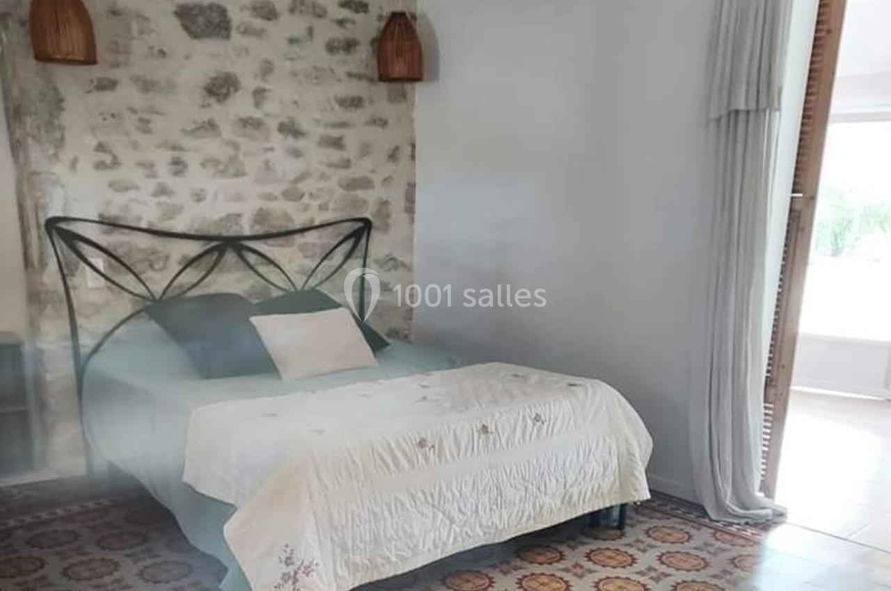 Lit double avec tête de lit en métal, draps clairs, mur en pierre apparente et sol en carreaux anciens.