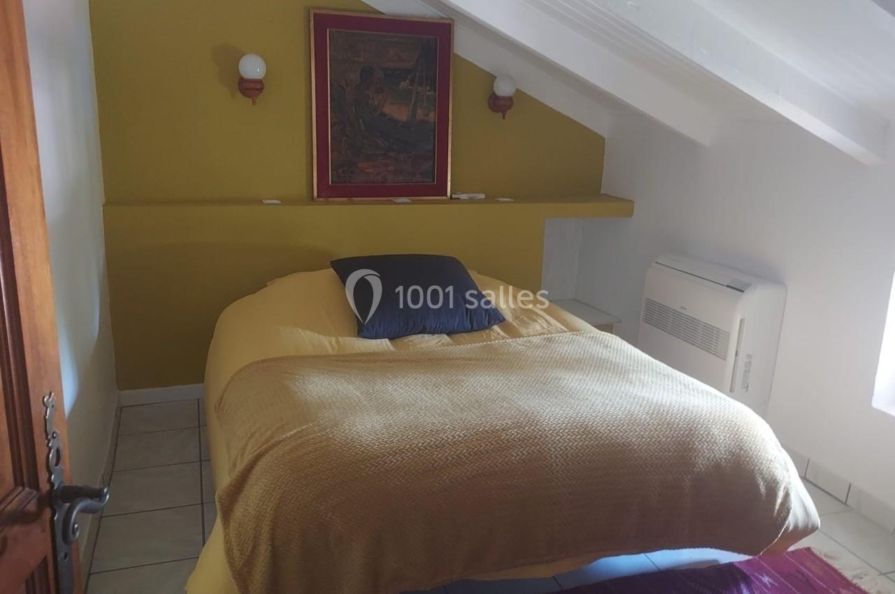 Chambre mansardée avec lit double, mur jaune, tableau encadré, deux appliques murales et lumière naturelle.