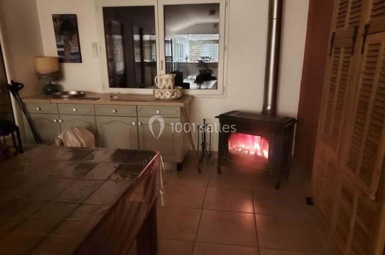 Poêle à bois allumé dans une pièce avec une table, un buffet, et une fenêtre donnant sur une autre pièce éclairée.