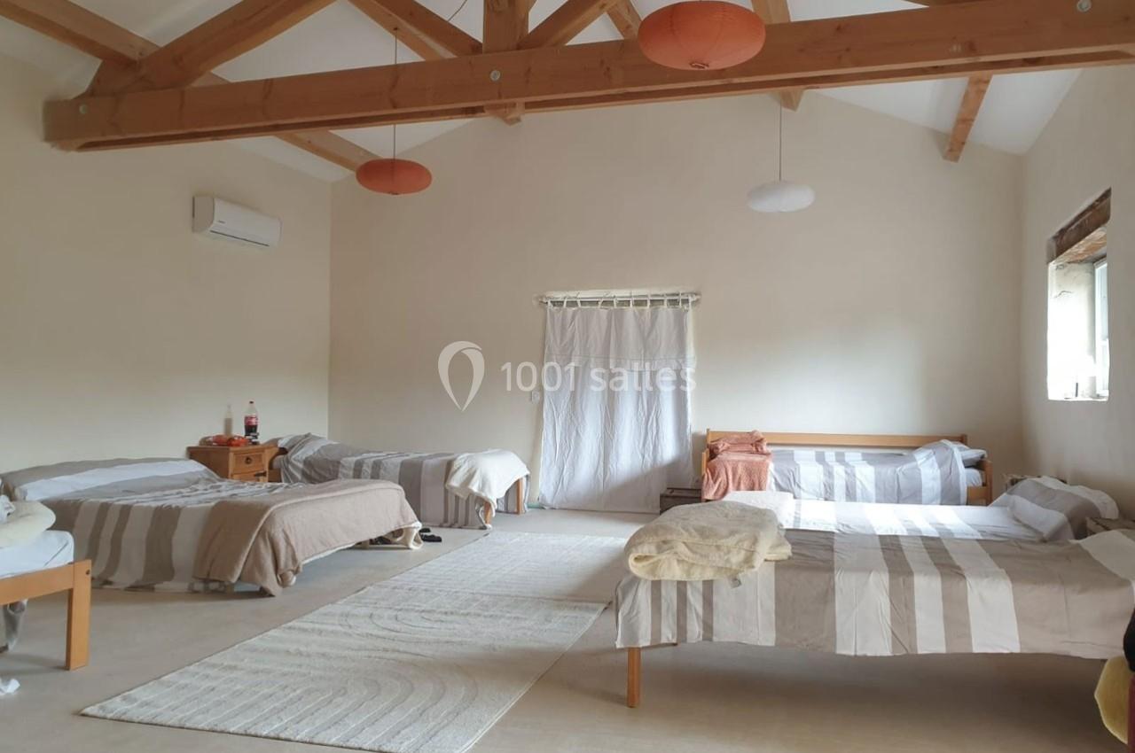 Chambre spacieuse avec quatre lits simples, poutres apparentes, éclairage suspendu et rideau blanc couvrant une porte.