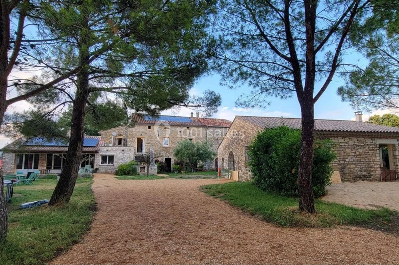 Cour d'une maison en pierre entourée de végétation, avec des arbres et un chemin en gravier au premier plan.