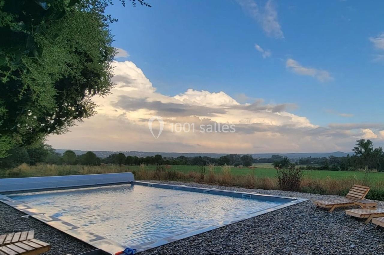 Piscine extérieure entourée de chaises longues, avec vue sur un paysage champêtre et un ciel partiellement nuageux.