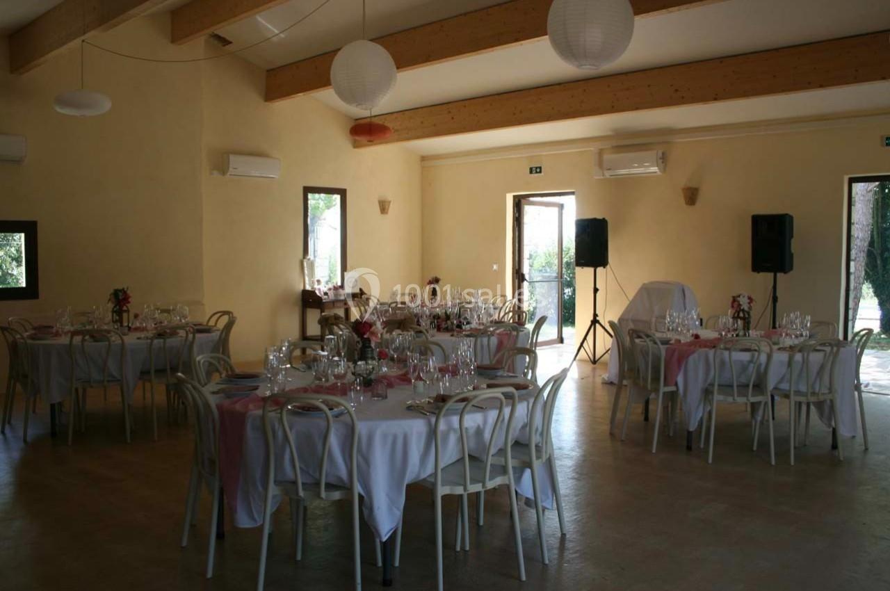 Salle de réception lumineuse avec tables rondes dressées, nappes blanches et décorations roses, sous un plafond à poutres…