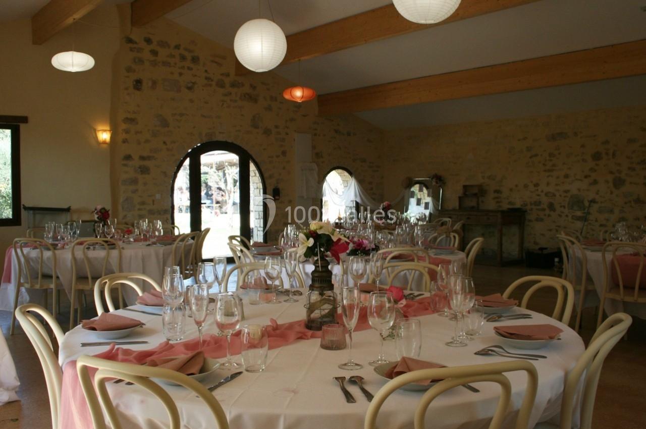 Salle de réception avec tables rondes dressées, nappes blanches, chemins de table roses et murs en pierre apparente.