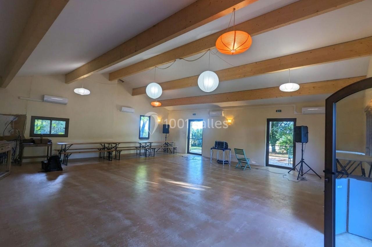 Salle lumineuse avec parquet, poutres apparentes, tables pliantes, chaises, éclairage suspendu et grandes fenêtres donnant…