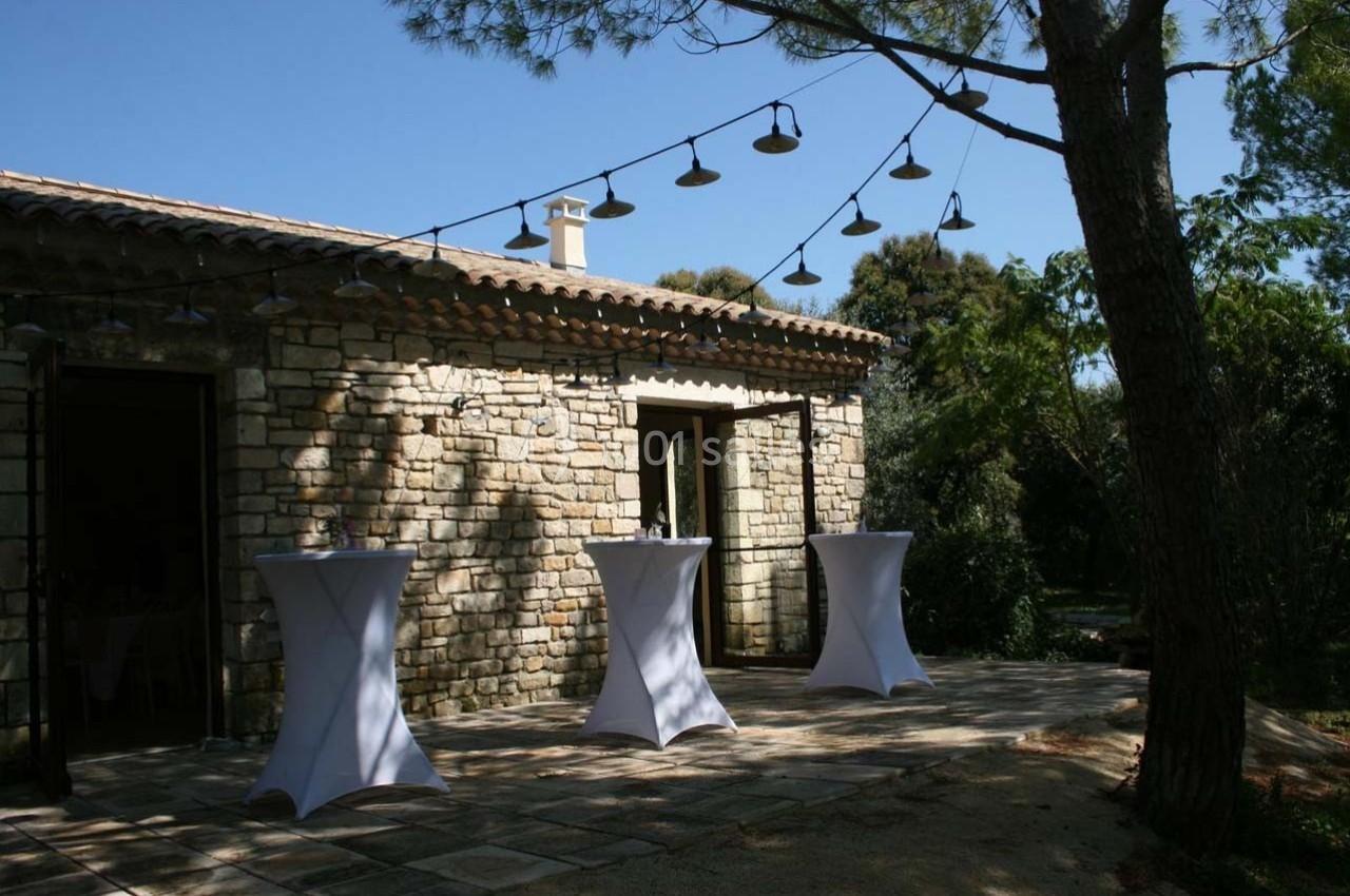 Terrasse en pierre avec tables hautes drapées de blanc, éclairée par des guirlandes lumineuses sous un ciel dégagé.
