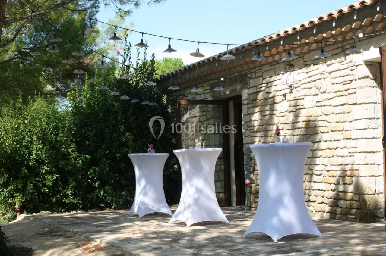 Terrasse en pierre avec trois tables hautes drapées de blanc, entourée de verdure et éclairée par des guirlandes lumineuses.