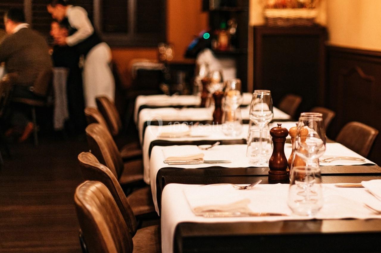 Salle de restaurant avec tables dressées, nappes blanches, verres et condiments, ambiance chaleureuse et tamisée.