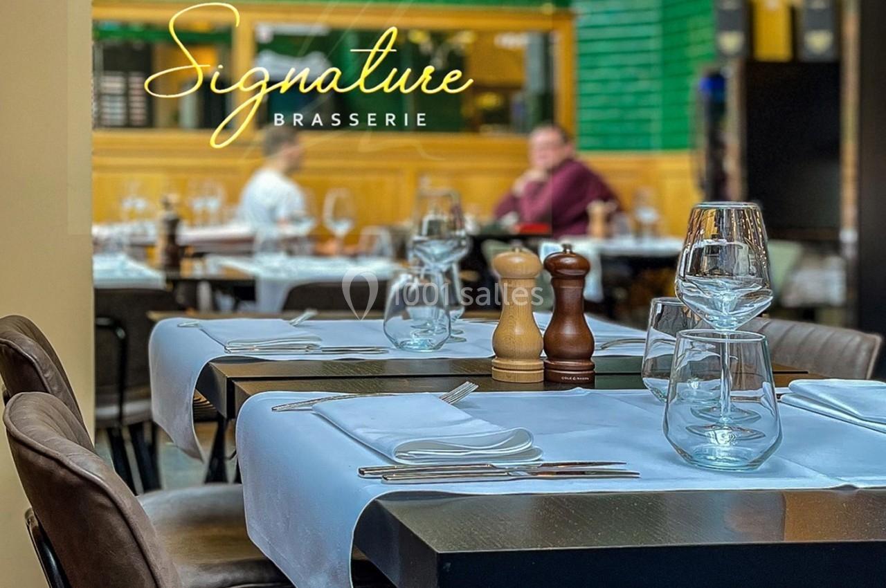 Table dressée avec nappes blanches, verres et couverts dans une brasserie avec des clients en arrière-plan flou.