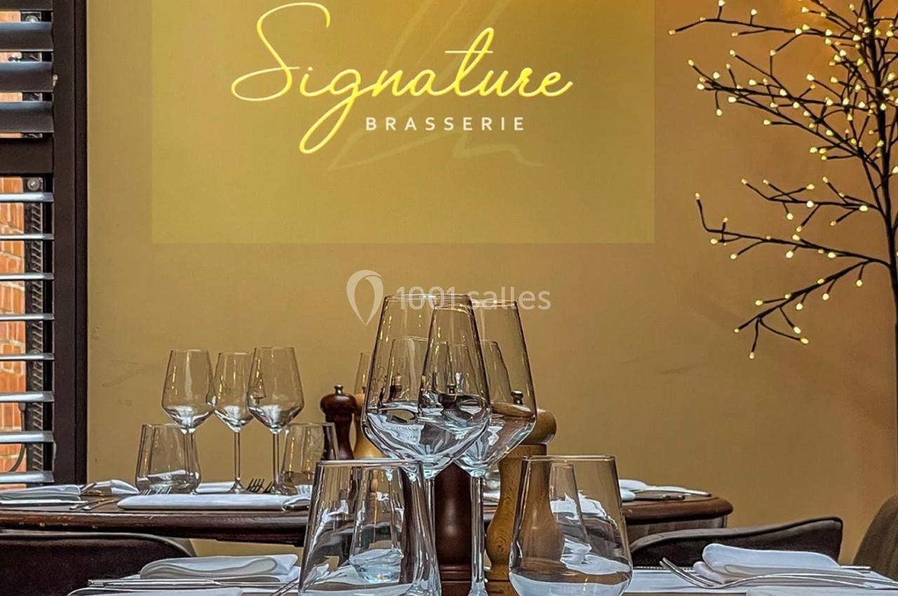 Salle de restaurant élégante avec tables dressées, verres et décor lumineux, logo ’Signature Brasserie’ en arrière-plan.