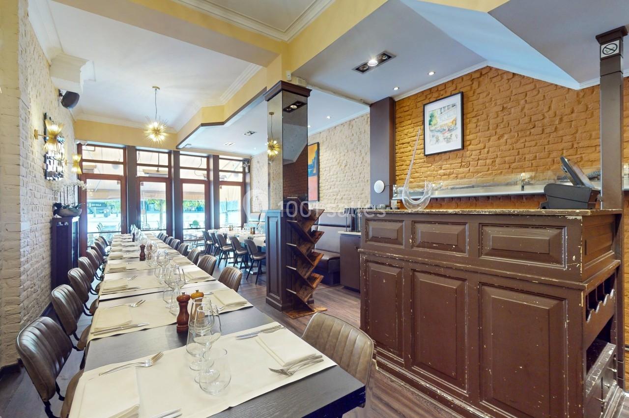 Salle de restaurant lumineuse avec tables alignées, nappes blanches, chaises en bois et comptoir en bois foncé.