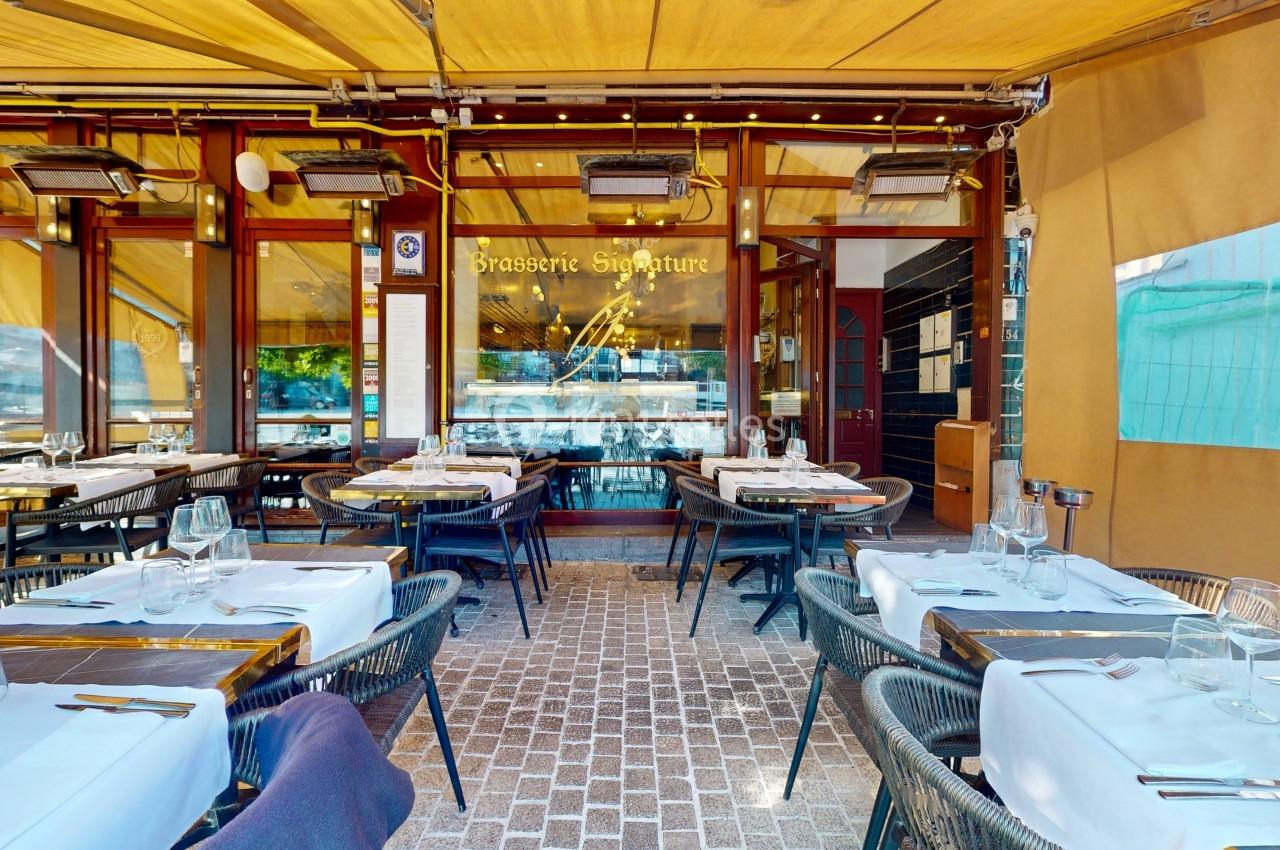 Terrasse d'une brasserie avec tables dressées, nappes blanches et enseigne visible sous un auvent jaune.