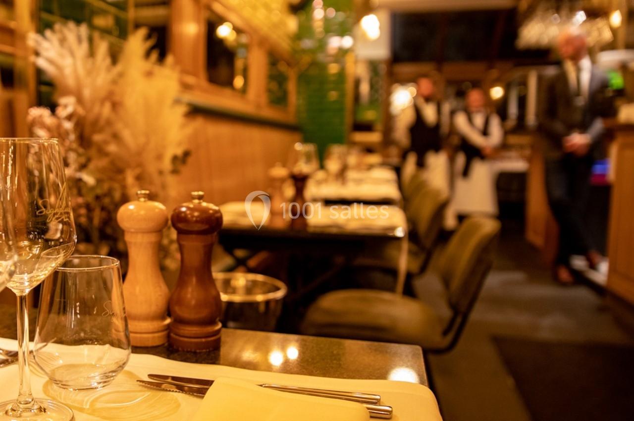 Salle de restaurant chaleureuse avec tables dressées, verres et couverts, éclairage tamisé et personnel en arrière-plan.