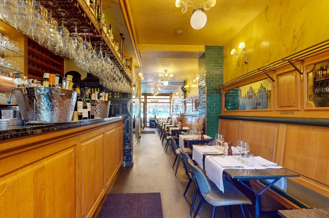 Salle de restaurant avec tables dressées, bar en bois à gauche, verres suspendus et éclairage chaleureux.