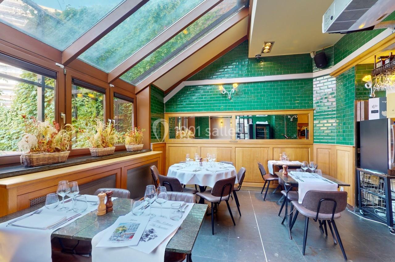 Salle de restaurant lumineuse avec verrière, murs en carreaux verts, tables dressées et plantes en décoration.