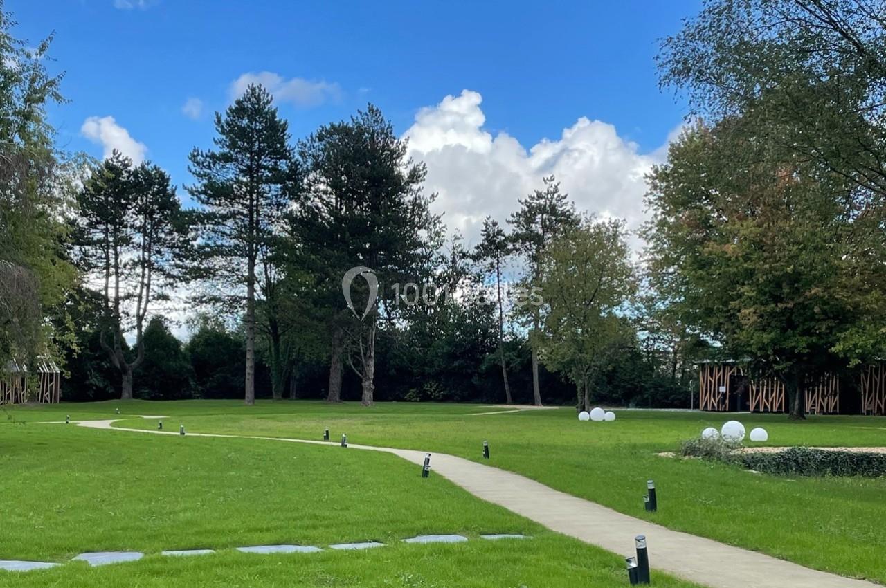 Allée pavée traversant un parc verdoyant avec des arbres, pelouse et éclairages extérieurs sous un ciel bleu.