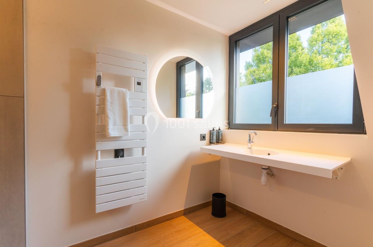 Salle de bain lumineuse avec un sèche-serviettes blanc, un miroir rond éclairé et un plan de travail minimaliste.