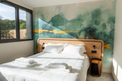 Lit avec des draps blancs, serviettes pliées au premier plan et mur décoré de branches et papillons colorés.