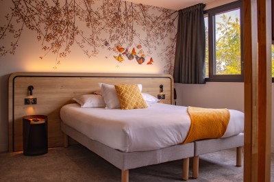 Lit avec des draps blancs, serviettes pliées au premier plan et mur décoré de branches et papillons colorés.