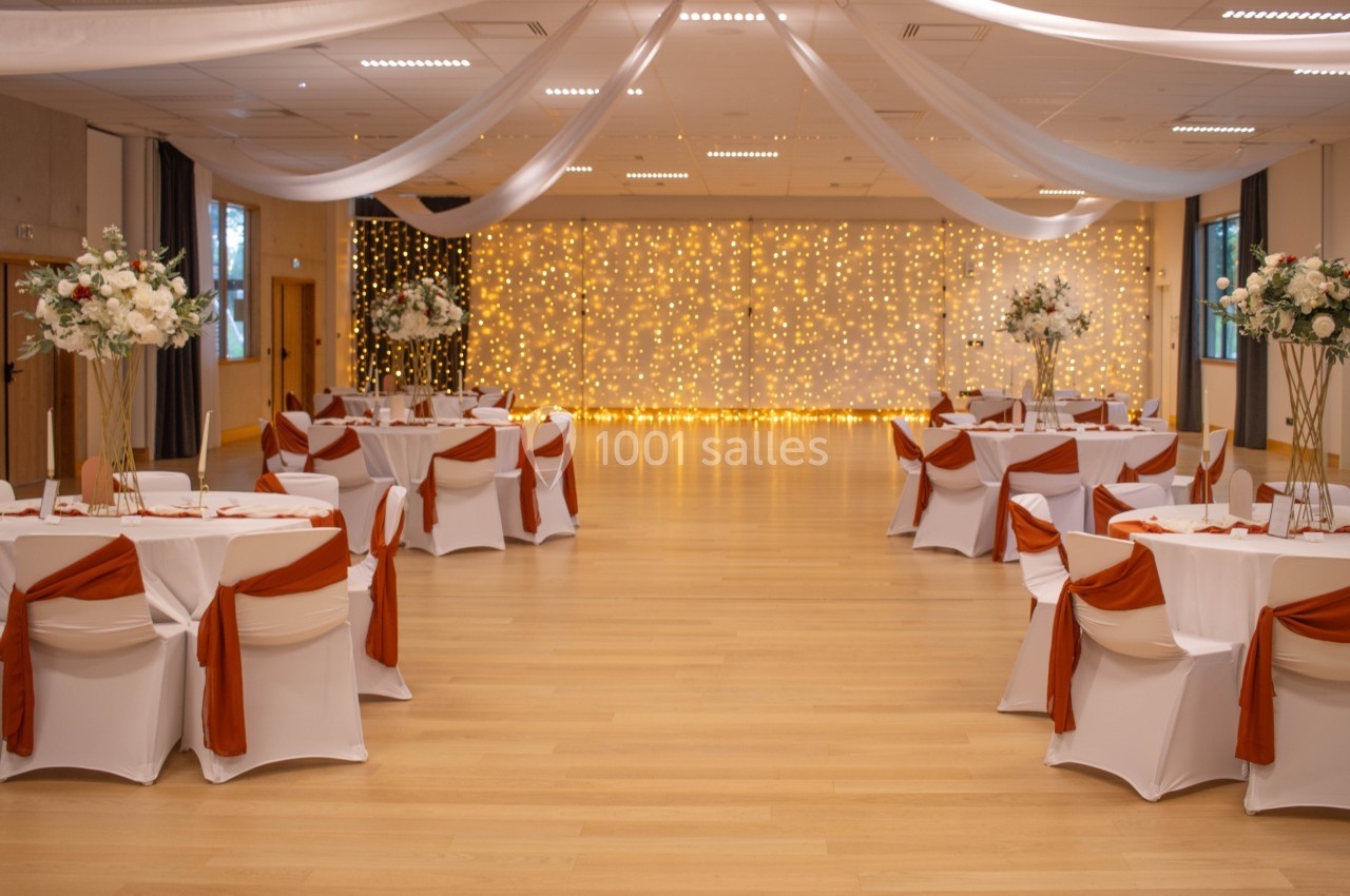 Salle de réception décorée avec des tables dressées, nappes blanches et rubans orange, éclairée par des guirlandes…