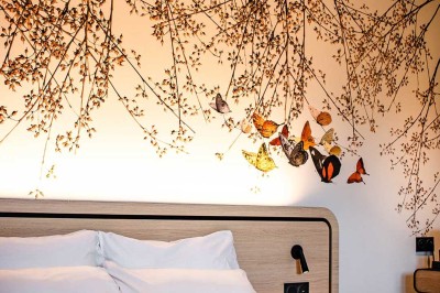 Lit avec des draps blancs, serviettes pliées au premier plan et mur décoré de branches et papillons colorés.