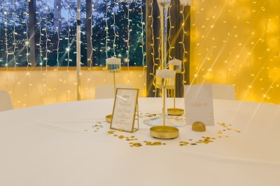 Miniature Location salle Liffré (Ille-et-Vilaine) - La Reposée #47 Table décorée avec des bougies, confettis dorés et un menu, devant une fenêtre ornée de guirlandes lumineuses.