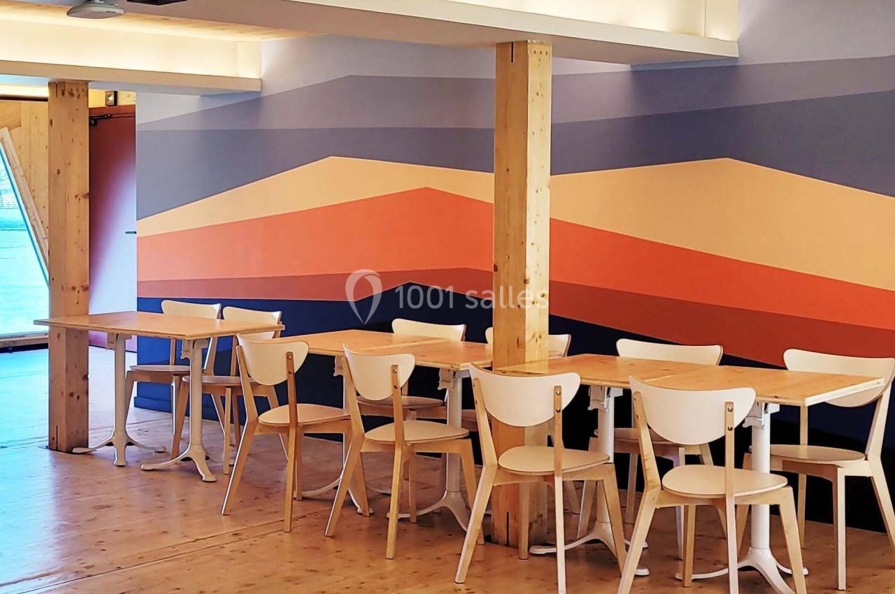 Salle lumineuse avec tables et chaises en bois clair, décorée d'une fresque murale aux motifs géométriques colorés.