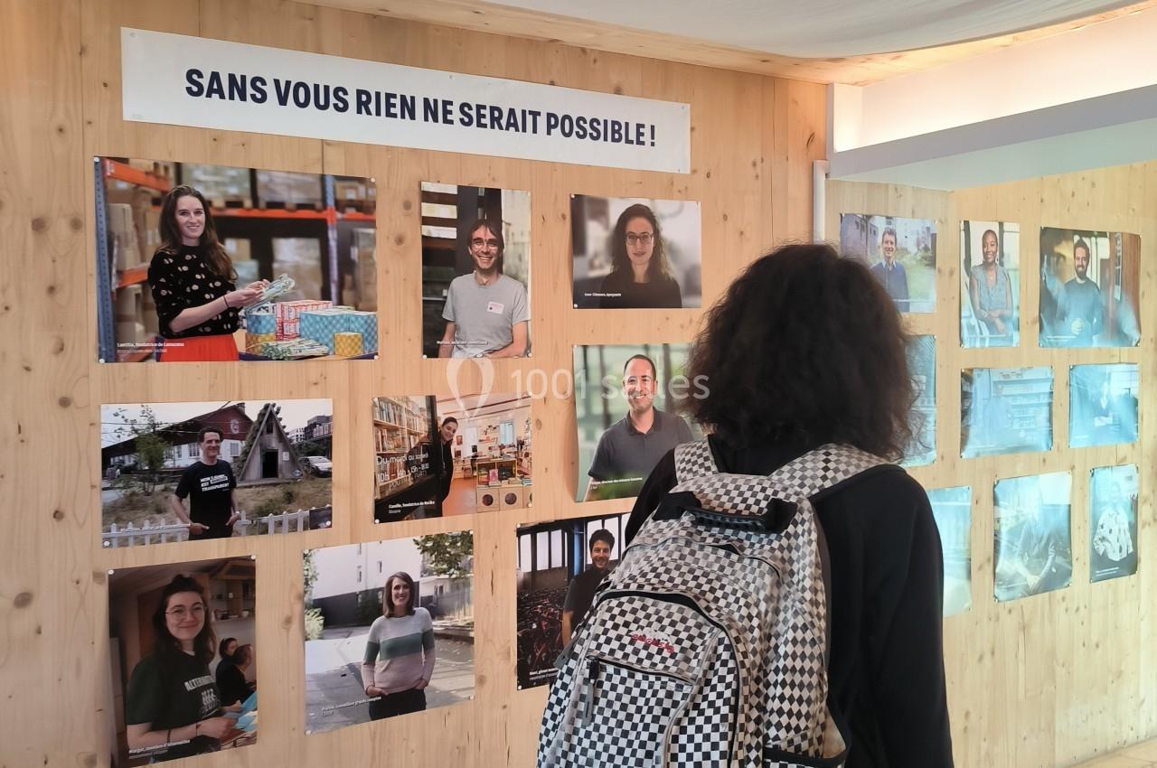 Une personne observe un mur d'exposition avec des portraits et un panneau indiquant ’Sans vous rien ne serait possible !’.