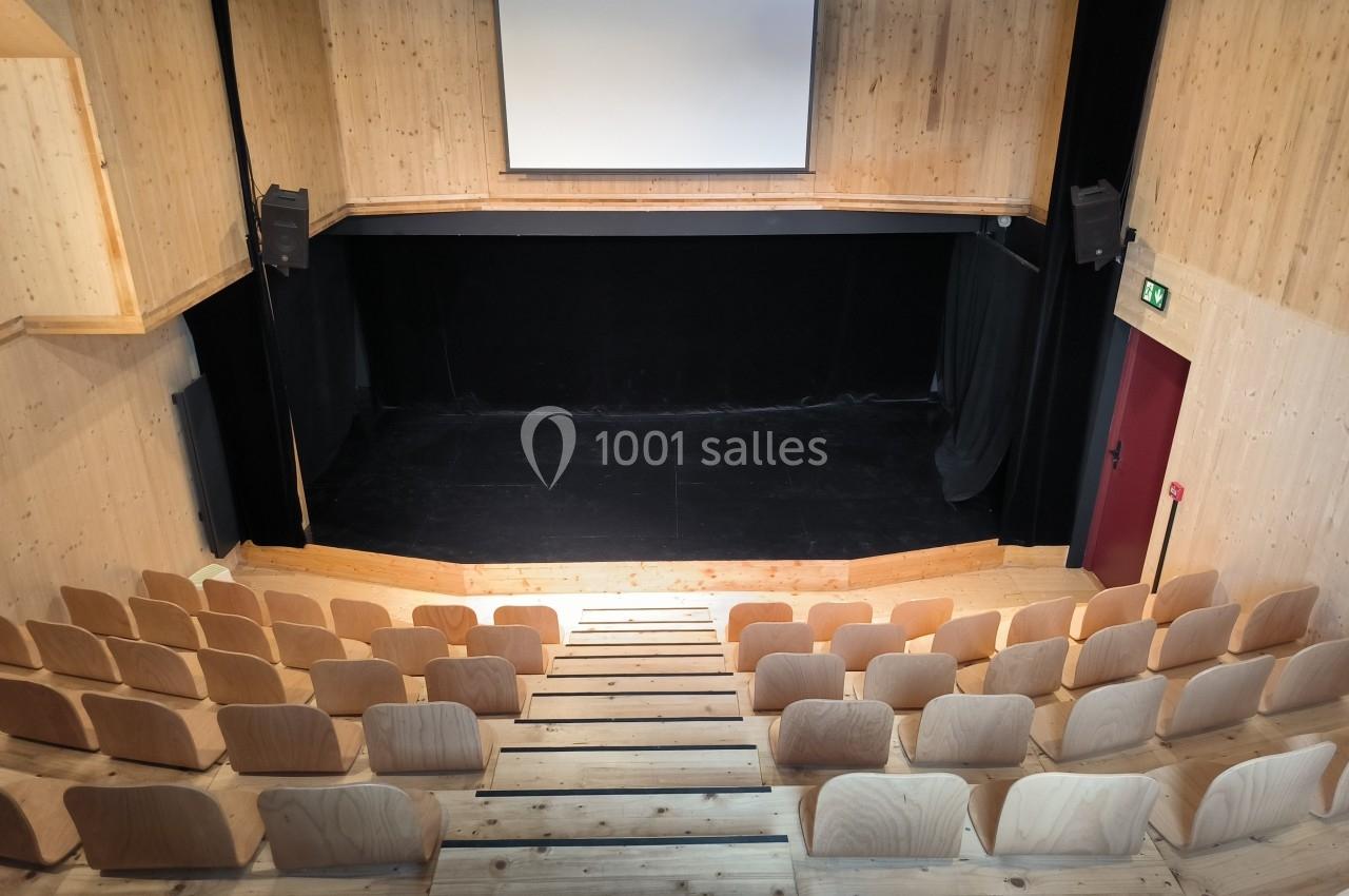Salle de spectacle en bois avec scène noire, écran blanc suspendu et rangées de sièges en gradins.